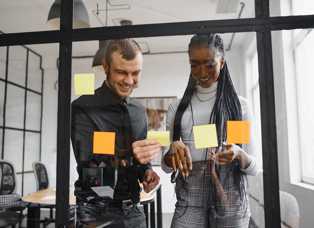 About Our Agency - Portrait Through the Glass of Two Cheerful Young Coworkers Adding Sticky Notes to the Glass During a Meeting