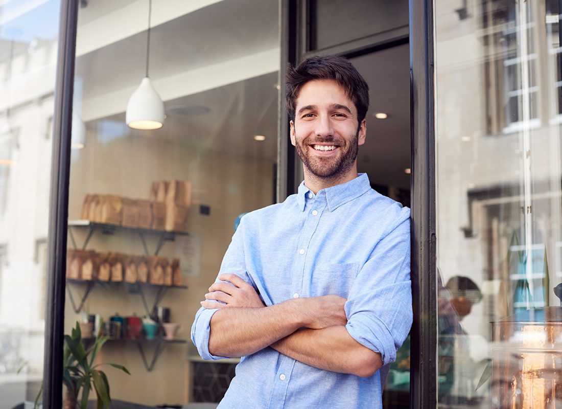 Insurance Solutions - Portrait of a Cheerful Young Business Owner Standing Outside the Front Door of his Main Street Shop with his Arms Folded